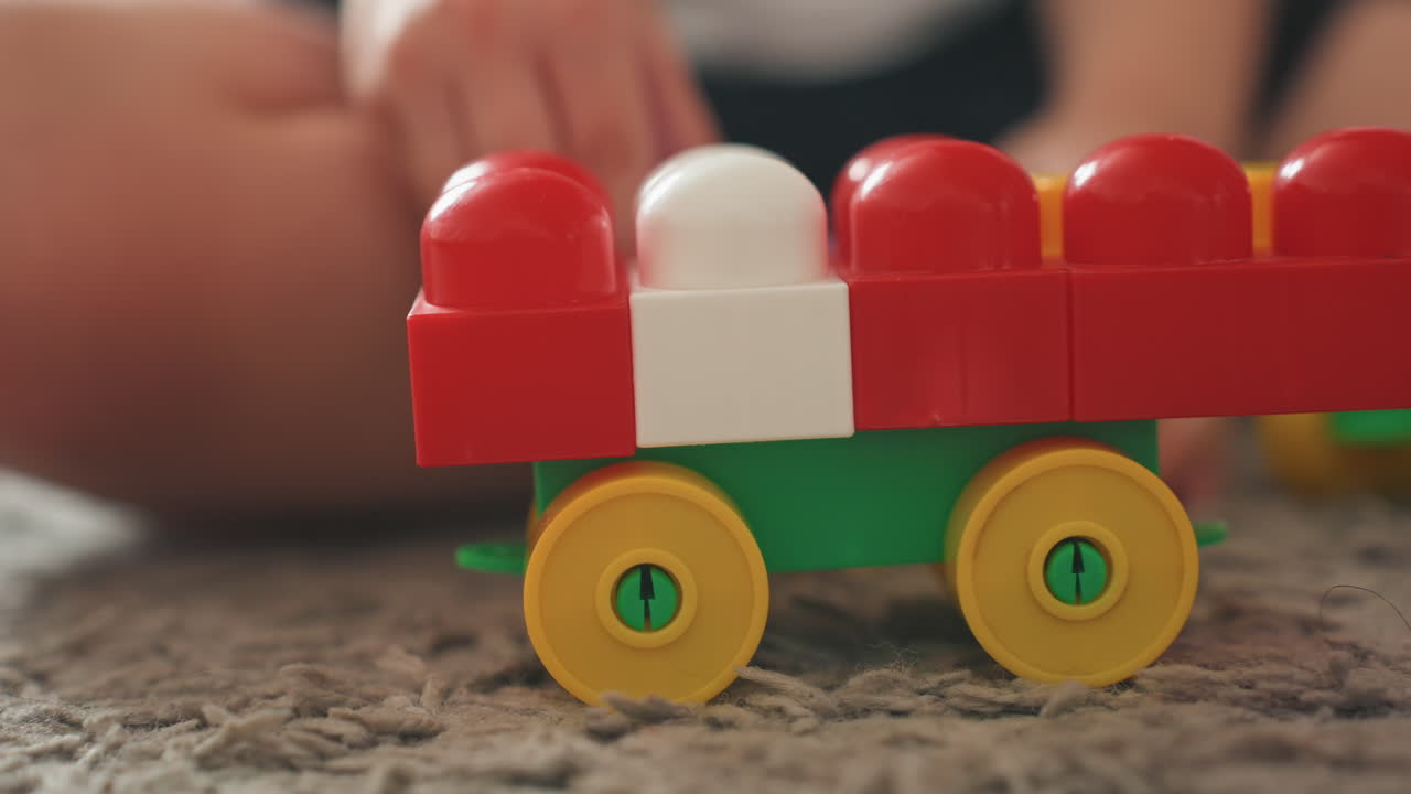 Hände eines Jungen schieben einen Spielzeugzug auf dem Teppich, Nahaufnahme von rollenden gelben Rädern und roten Plastikbausteinen, taktiles Spiel und Feinmotorikübung, gemütliche Szene auf dem Boden im Innenbereich mit warmem Naturlicht und spielerischer Atmosphäre.