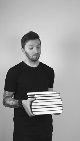 grayscale footage of a tattooed man with short hair and a trimmed beard lifting up a big pile of hardcover books and shaking his head showing that he is not pleased or happy with the amount of books.