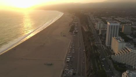 High-up-view-from-Above-over-Pacific-Coast-Highway-PCH-next-to-Santa-Monica-Beach-Ocean-Waves-in-Los-Angeles-with-light-traffic-at-Golden-Hour-Sunset,-Aerial-establisher