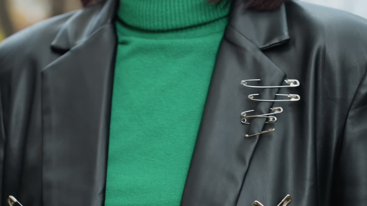 primo piano di modella con dolcevita verde e giacca di pelle con spille su sfondo naturale all'aperto, indossa un blazer di pelle nera sopra una vivace maglia verde, spille da balia raggruppate a forma di spilla sul risvolto, look cool