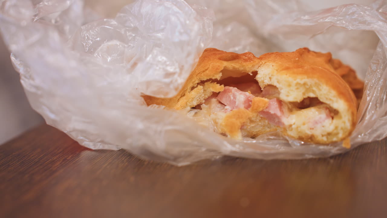Dolce per la colazione avvolto nella plastica sul tavolo, un panino croccante ripieno di uovo, prosciutto e formaggio adagiato in una sottile pellicola trasparente su un bancone di legno. La vista ravvicinata mostra briciole, la consistenza e un aspetto unto, evocando un pasto veloce.