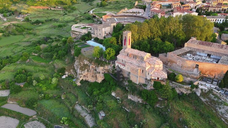 Drone flies backward and upward over the Romanesque church of Brihuega, Spain, revealing the cliff, cemetery, bullring, and village under golden sunrise light. Historic and scenic architectural view