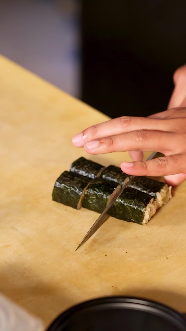 Detailní záběr rukou šéfkuchaře precizně krájících sushi rolky obalené mořskou řasou na dřevěném prkénku