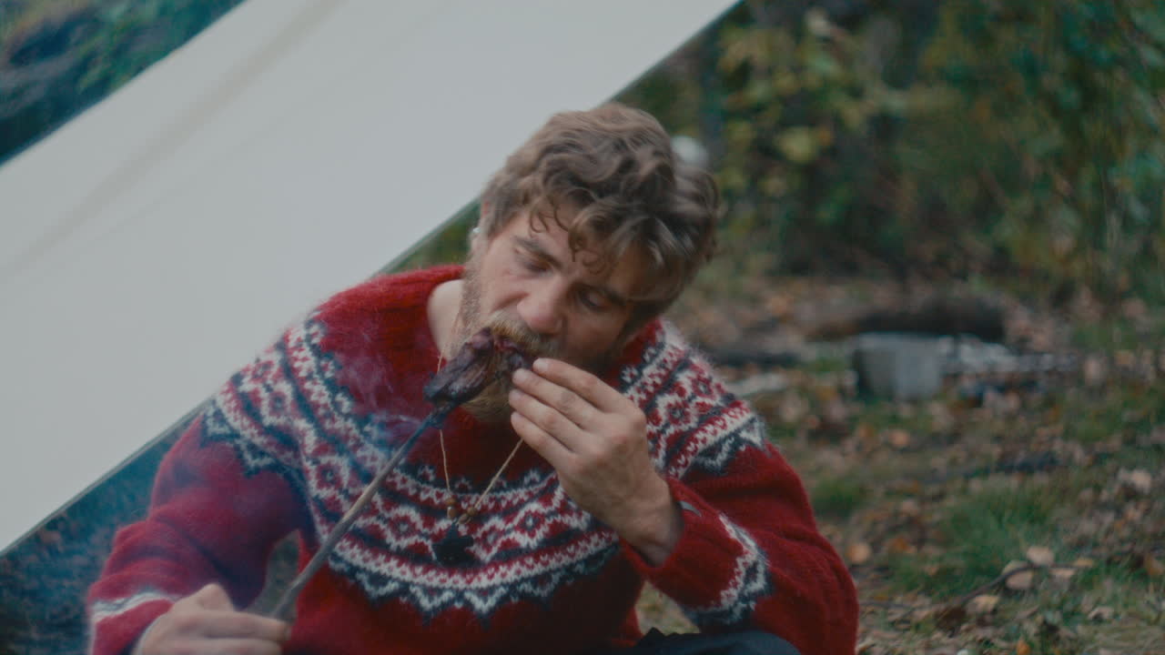 Man Eating Roasted Meat on Stick by Campfire and Tent in Forest