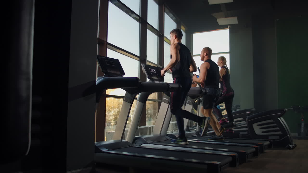 Two men running on a treadmill in the background of a large window in the fitness room. Young men running in the fitness room. Aerobic workouts in the gym.