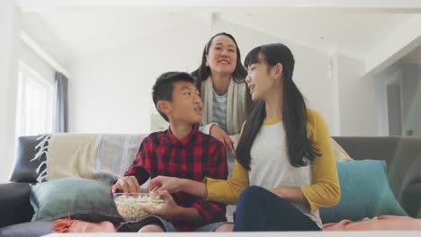 happy asian mother in living room watching tv with son and daughter sitting on couch eating popcorn