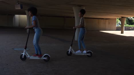 Two-mixed-race-women-riding-electric-scooter-under-bridge