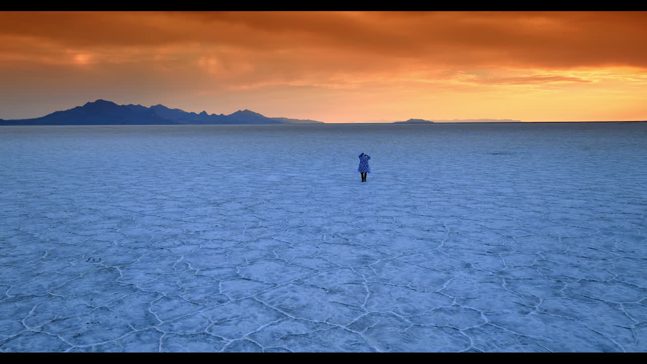 Catching up with a man wearing a coat and walking by the salty plain. Mountain silhouettes and orange sky at backdrop. Bonneville Salt Flats, Utah, USA at sunset time