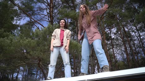 two young girl friends make fools of themselves on top of the roof of a caravan in the middle of the forest looking at the camera.