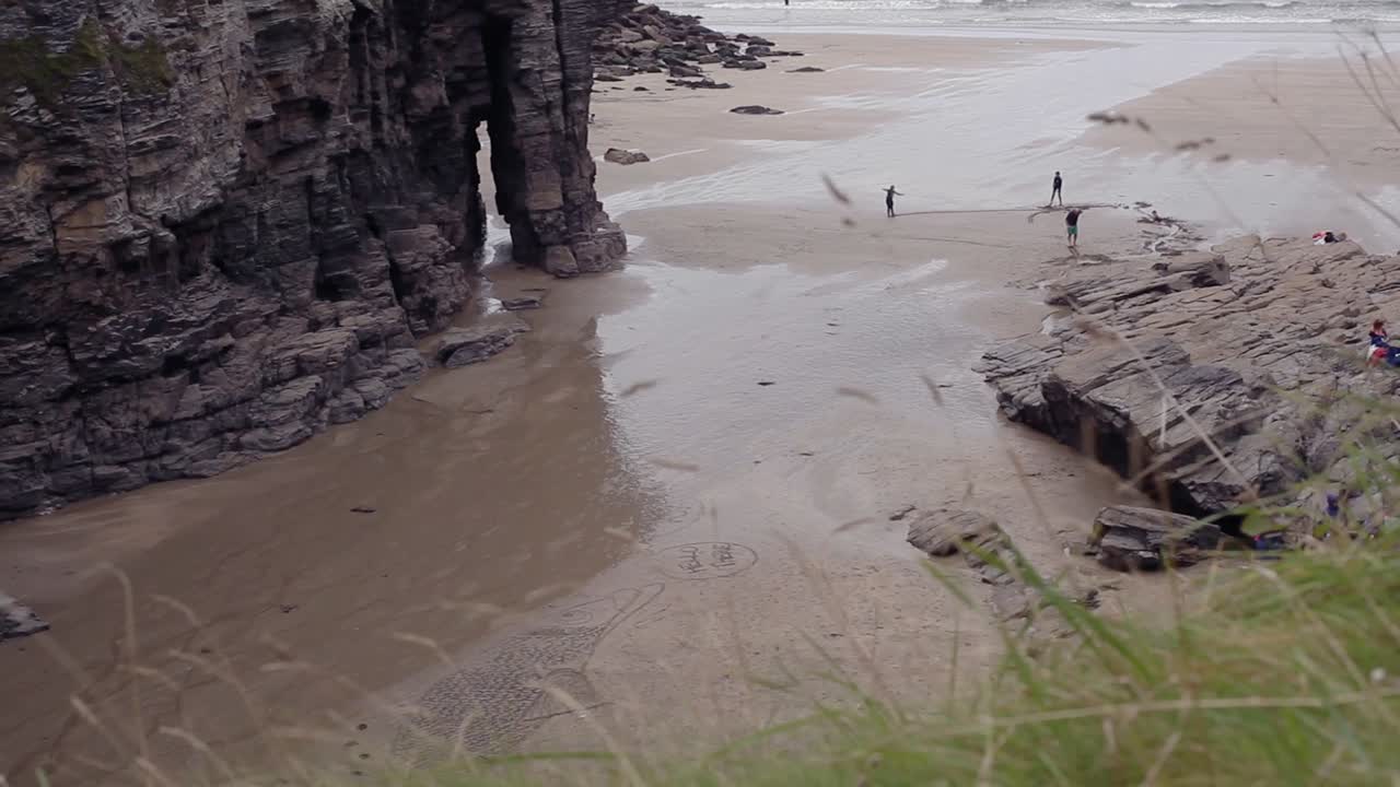 Summer day in a Cornish sandy bay. Holiday makers enjoy there time and draw something in sand. Rocky mountain is in sight and green grass slightly blurred sways in foreground. Footage in HD