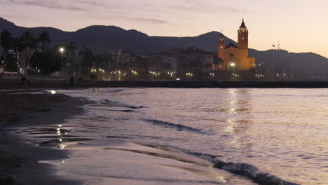 coastal sunrise skyline is reflected in sea waves washing ashore, seagulls fly by as silhouette of church stands against backdrop of majestic mountain range