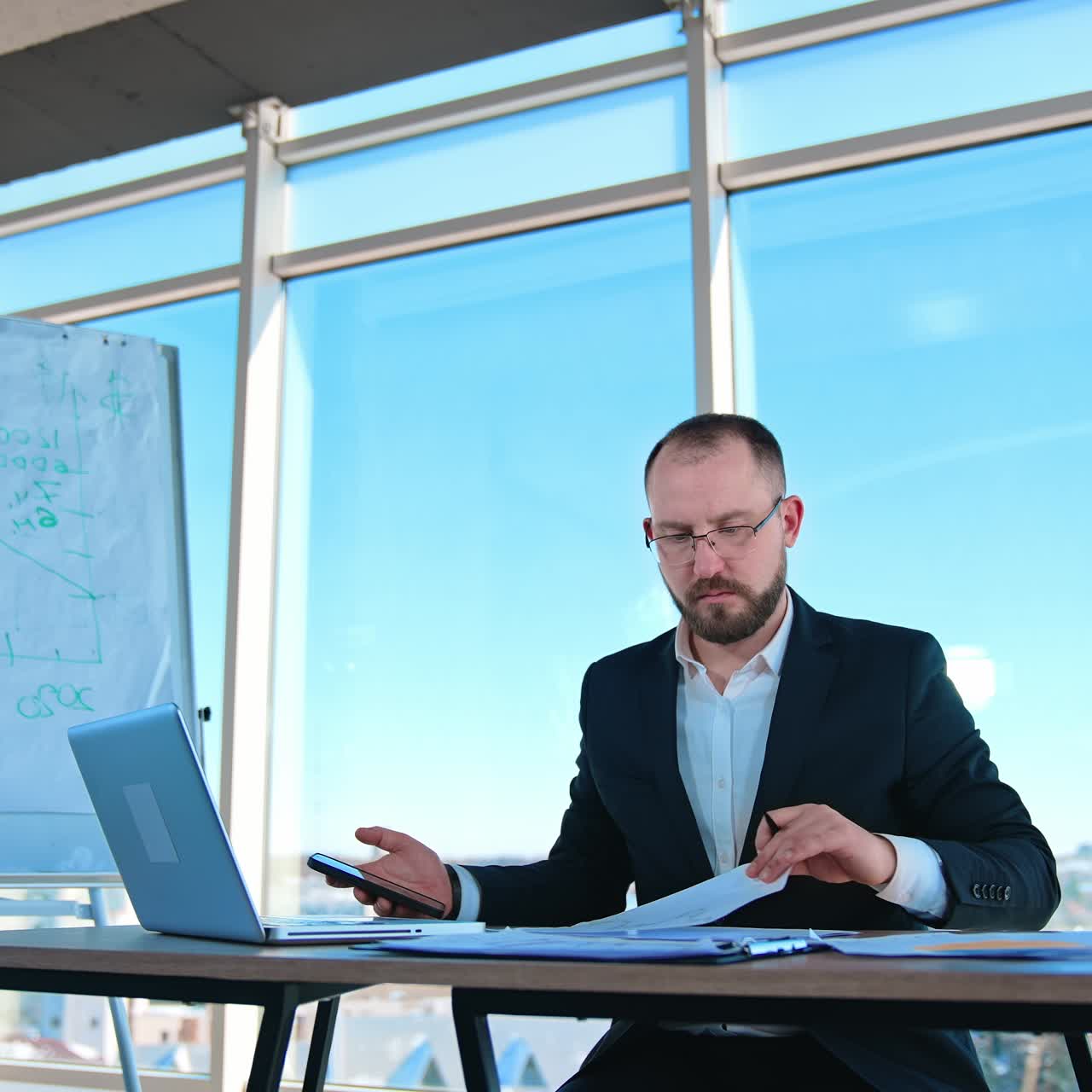 Focused businessman uses smartphone in workplace. Bearded entrepreneur in suit and glasses sitting at table and working next to large window