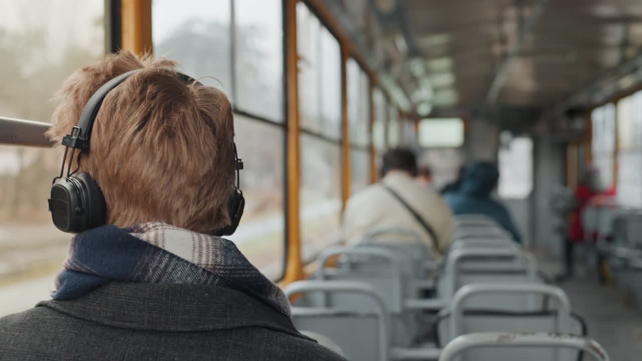 Weißer Mann im Bus hört zu, Podcast-Kreativer nimmt Klänge auf, Rückenansicht mit Kopfhörern und Schal, Pendler im Mantel beobachtet bewölkte Vorstadtlandschaft durchs Fenster, nachdenkliche Einsamkeit