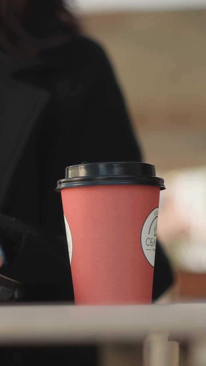 Close-up of coffee cup on table with person sitting, partially obscured by laptop edge, indoor workspace setting featuring blurred background