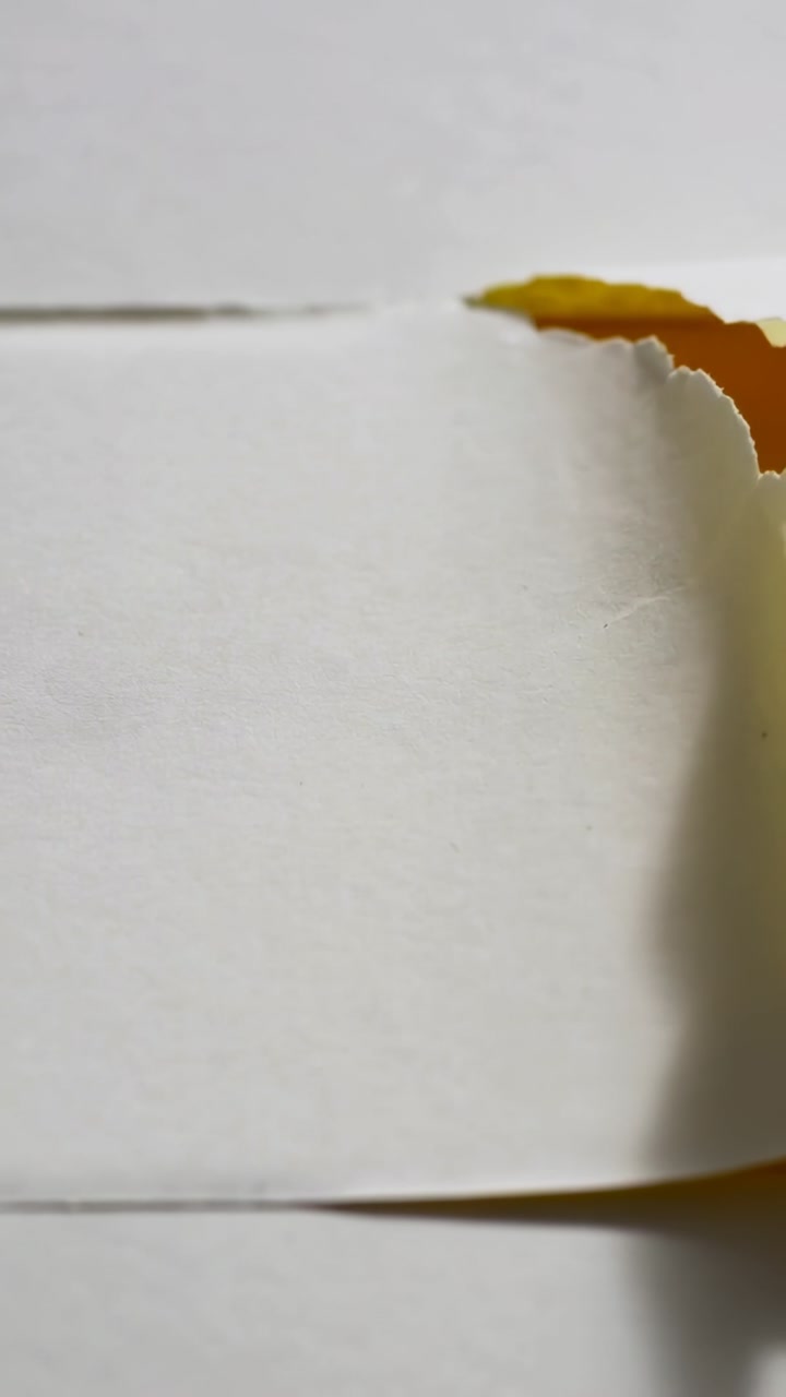 Vertical video: Starting small tear tearing white sheet, revealing yellow layer on flat surface