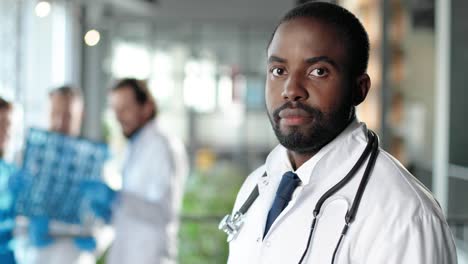 portrait of african-american physician in white gown looking at camera, smiling and standing in clinic