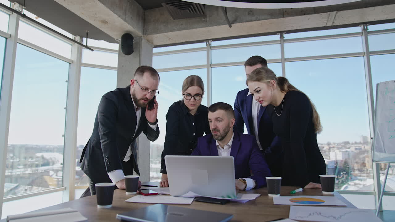 Collaborative team crowded together around their ceo in office. Man in center at computer proposes ideas to discuss. One employee answers the phone. Close up with gradual distancing.