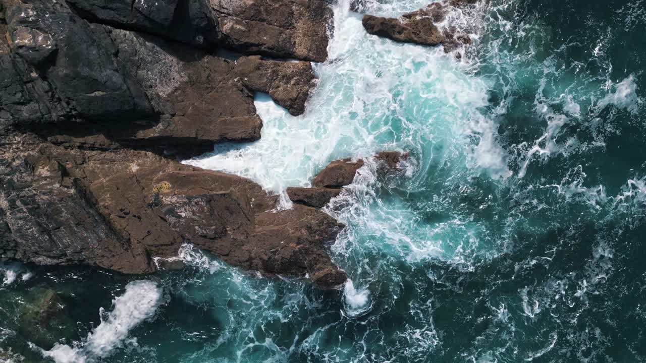 Slow-motion birdseye view of foamy waves hitting rocky shoreline in Cornwall, England, natural backdrop background
