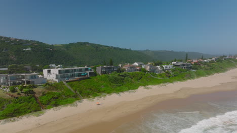 Fly-along-row-of-vacation-houses-on-sea-coast.-Waves-washing-narrow-sand-beach-on-sunny-day.-South-Africa