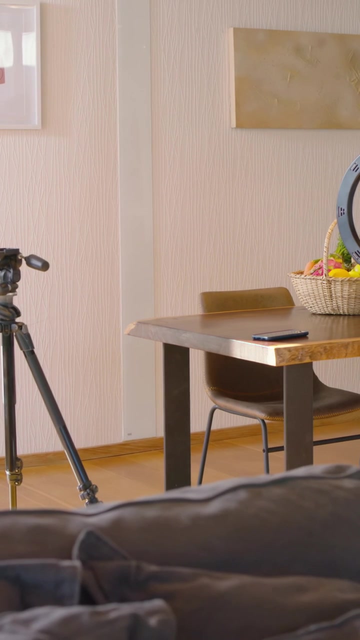 Verticale video: Vrouw in witte top en korte broek komt binnen, stelt ringlicht bij aan tafel en bereidt opname voor.