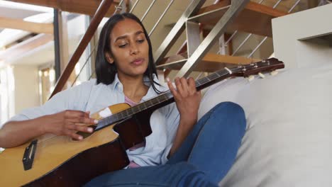 mujer de raza mixta en el sofá en casa tocando la guitarra y cantando