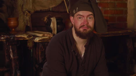 a man dressed in medieval clothing is standing in a dimly lit room, looking at the camera. he has a beard and is wearing a hat. the room is decorated in a rustic style, with wooden furniture and a candle on the table.