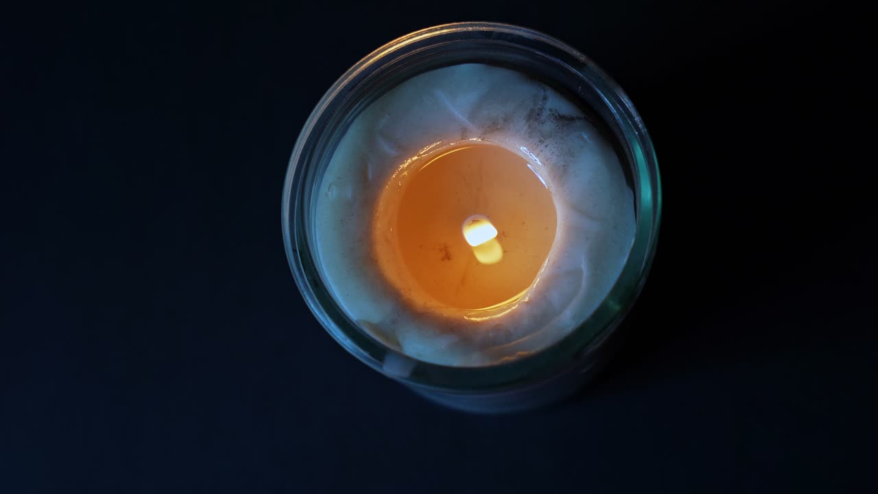 Primo piano dall'alto di una candela accesa in vasetto con fiamma tremolante e cera fusa, che brilla caldamente contro uno sfondo scuro di notte, creando un'atmosfera calma e accogliente.