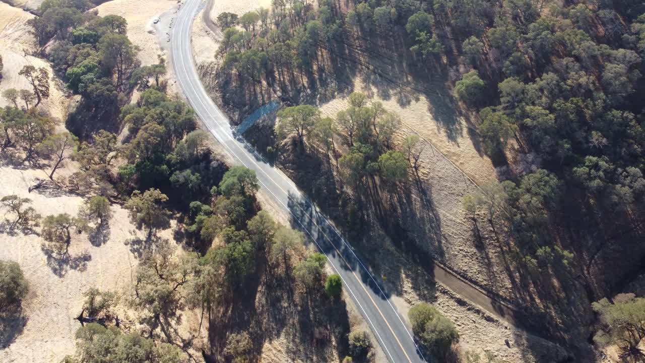 Traveling drone moving along isolated winding road lined with trees and golden hills