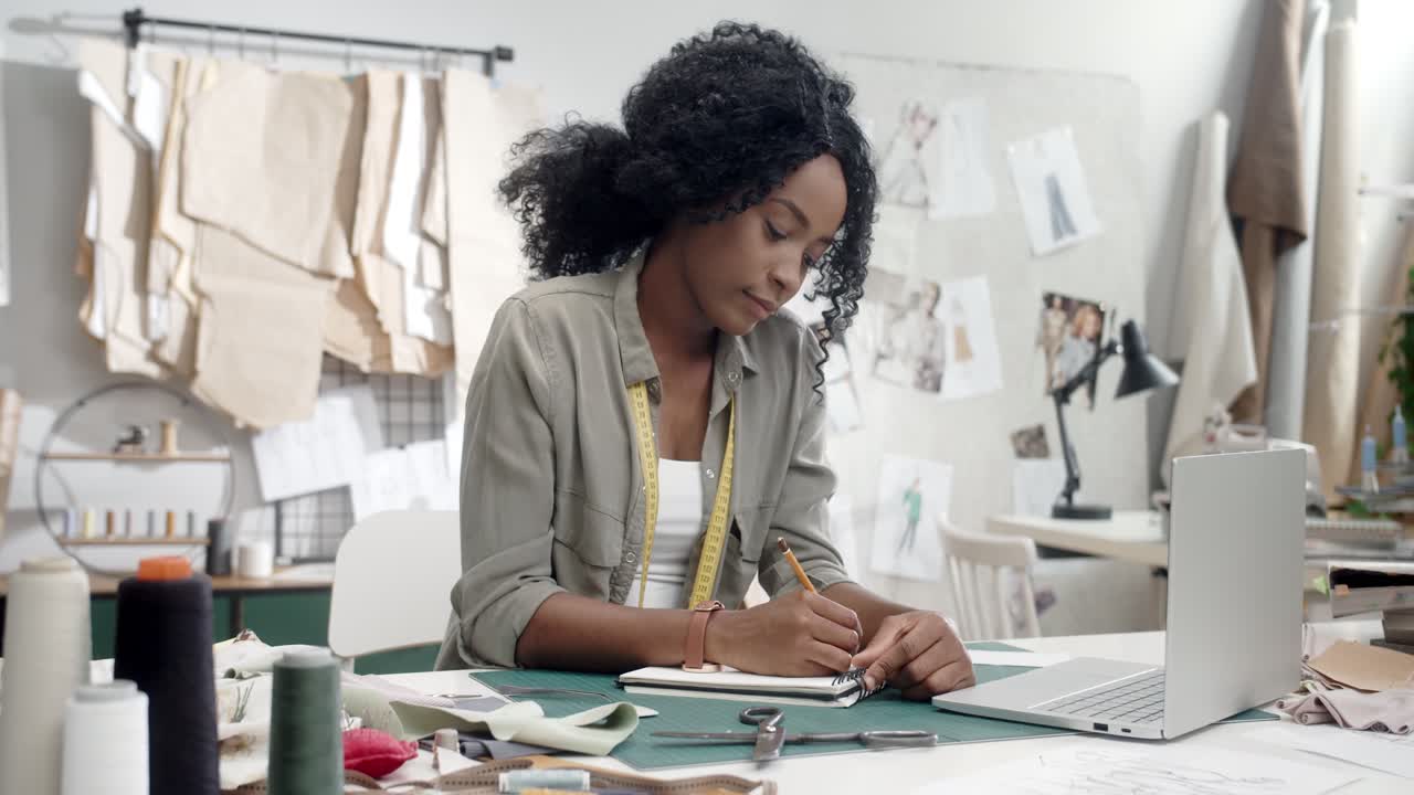 Woman Clothes Designer Sitting And Working At The Laptop Computer, Drawing Outlines In Her Notebook And Smiling To The Camera In Her Studio 1