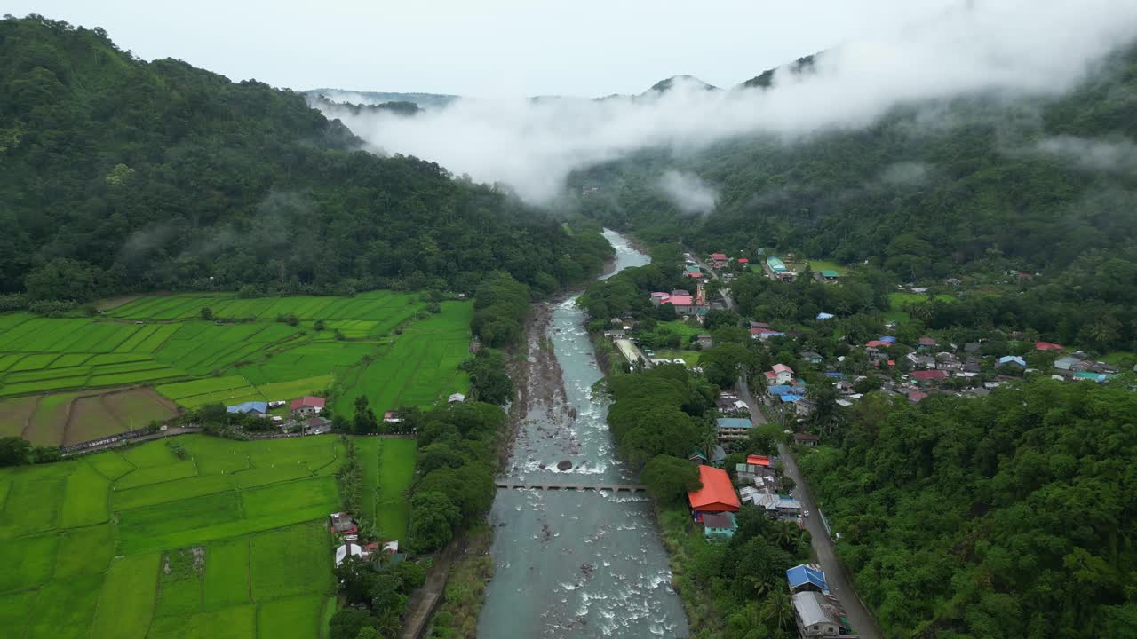 A pull-out aerial shot starts over rice paddies and riverside homes, then widens to reveal forested hills and a misty Philippine valley with vibrant village rooftops