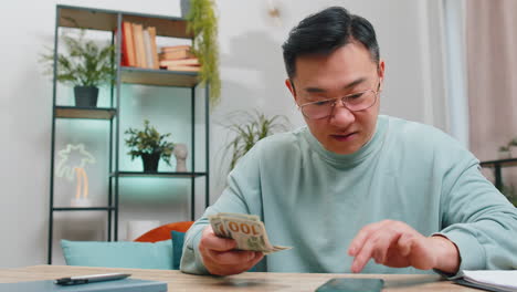 Rich happy asian man counting money cash on smartphone calculator app calculate income earnings