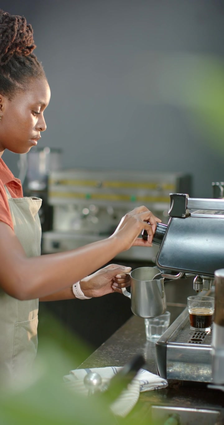 Vertical video: Woman in apron using steam knob and frothing milk pitcher for latte at cafe counter