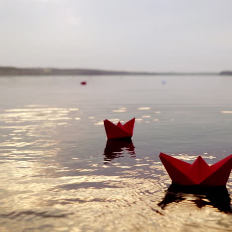 woman's hand is putting red paper boat on the water to others swimming at sunset. origami ships on waves on river in the evening
