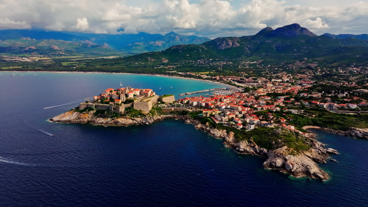Aerial drone shot over the historic old town of Calvi, Corsica, France. Picturesque view of coastal town, landscape, fluffy clouds, and mountains in the background
