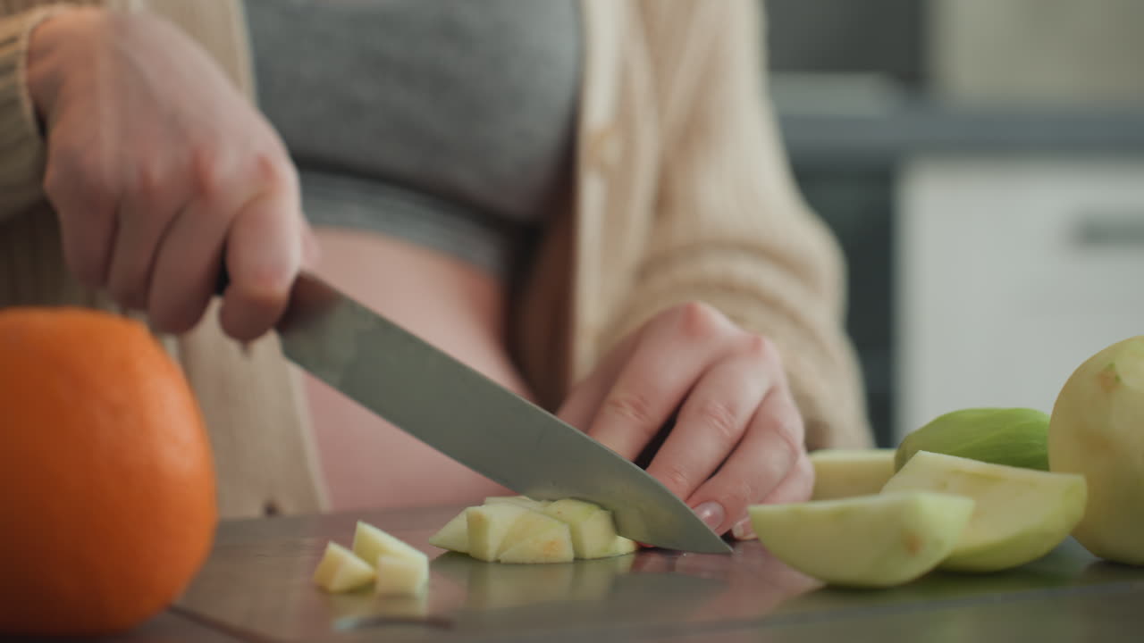 Pregnant woman slicing apple on countertop, quiet morning scene with expectant mother preparing vitaminrich fruit snack, closeup hands chopping apple beside orange, soft natural light, cozy cardigan