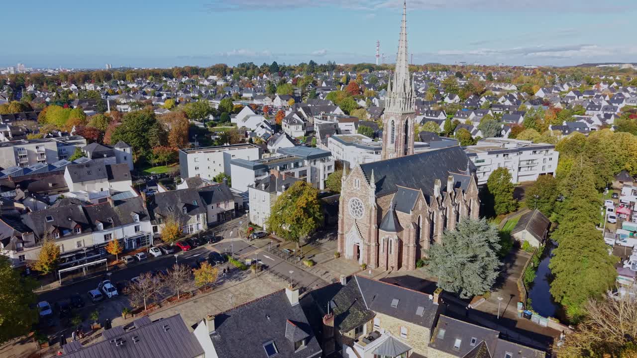 Saint-Martin Church in Cesson-Sévigné, Brittany, France, surrounded by residential houses, trees with autumn colors and modern buildings under clear daylight skies, drone establishing shot