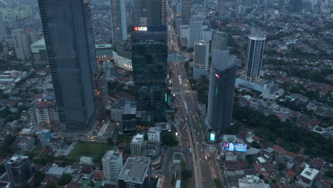 Toma-Aérea-De-Vehículos-En-Una-Concurrida-Carretera-De-Varios-Carriles-Y-Rotonda-Al-Atardecer-En-El-Moderno-Centro-De-La-Ciudad-De-Yakarta,-Indonesia