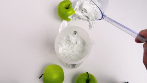 Birdseye-static-shot-from-a-shaker-filled-with-water-while-adding-a-scoop-of-finest-green-apple-whey-powder-for-a-tasty-shake-with-green-apples-on-the-side