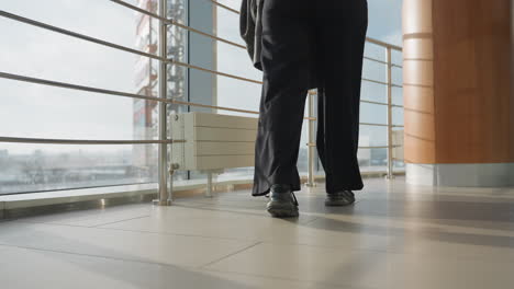 low angle rear view of person in black pants and sneakers walking across tiled indoor floor while holding jacket, surrounded by modern railings and large glass windows in contemporary urban space