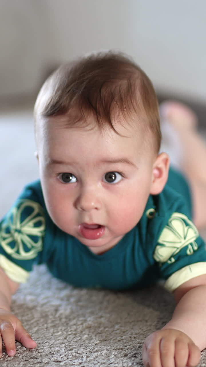 Sweet Caucasian baby boy lies on the carpet banging hands over the floor. Cute infant child holds still a little looking at camera attentively. Vertical video
