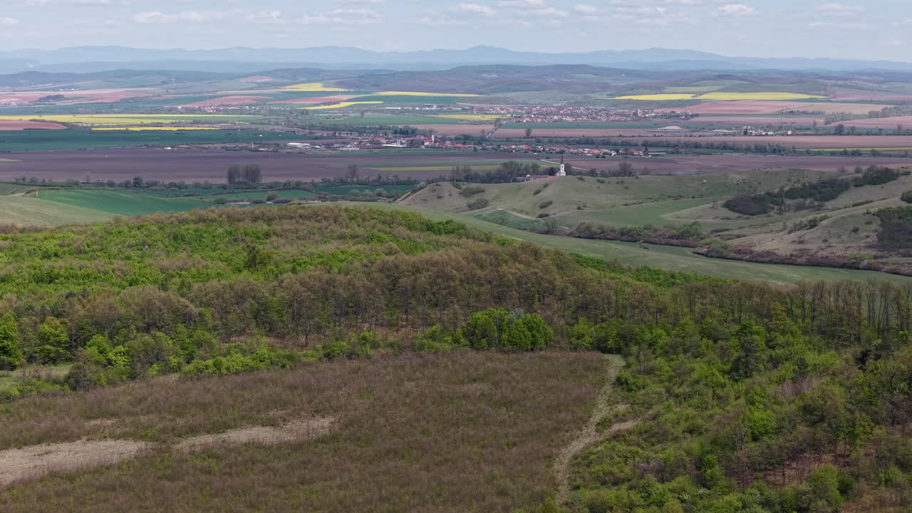 Een pittoreske hoge-hoekopname legt een sereen landelijk landschap vast, met glooiende groene heuvels, bossen en gecultiveerde velden, leidend naar een ver dorp en bergen aan de horizon.