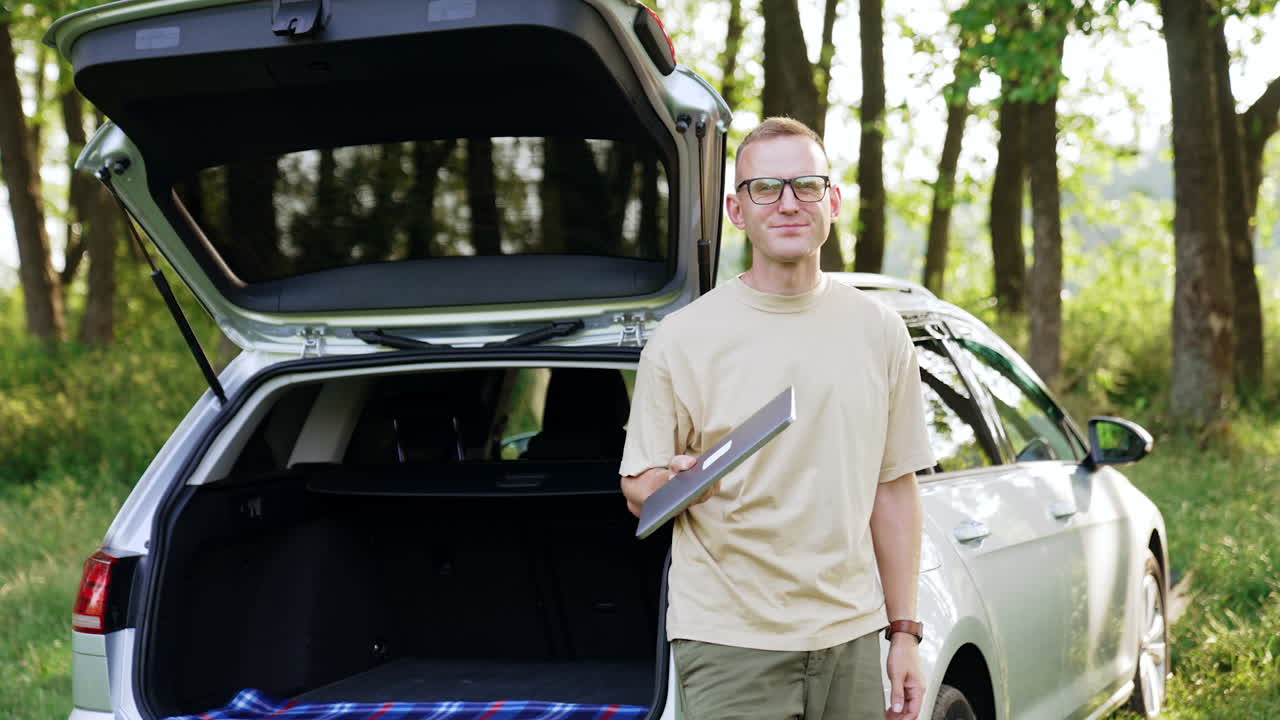 Serious Caucasian man holding a laptop in hands standing near the car outdoors. Male opens gadget and switches it on starting work.