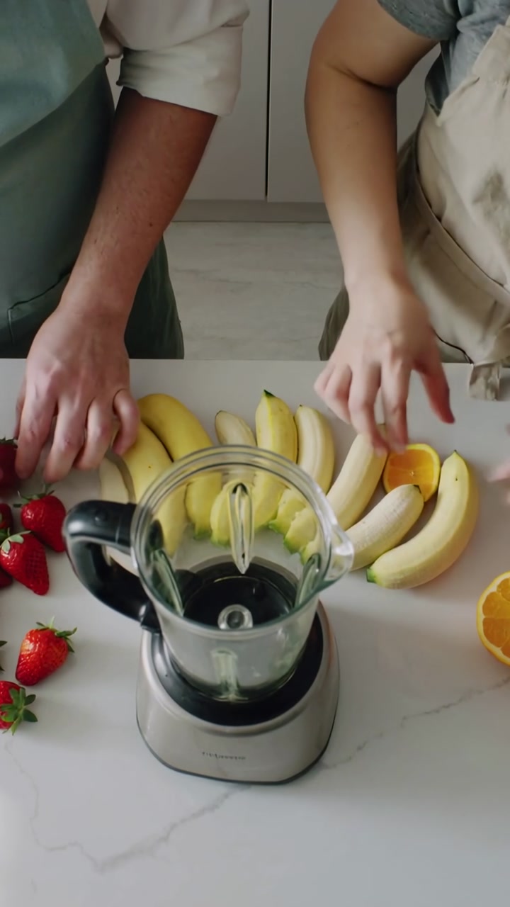 Vertical video: Starting smoothie prep adults arranging fruit at kitchen counter, blender, aprons