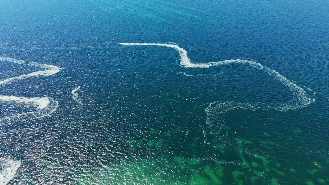 Jet ski racing takes place on a bright sunny day in clear water, with riders navigating through twisting paths on the shoreline