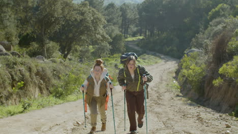 front view of two young female hikers walking up wide dirt road with trekking poles on bright autumn day