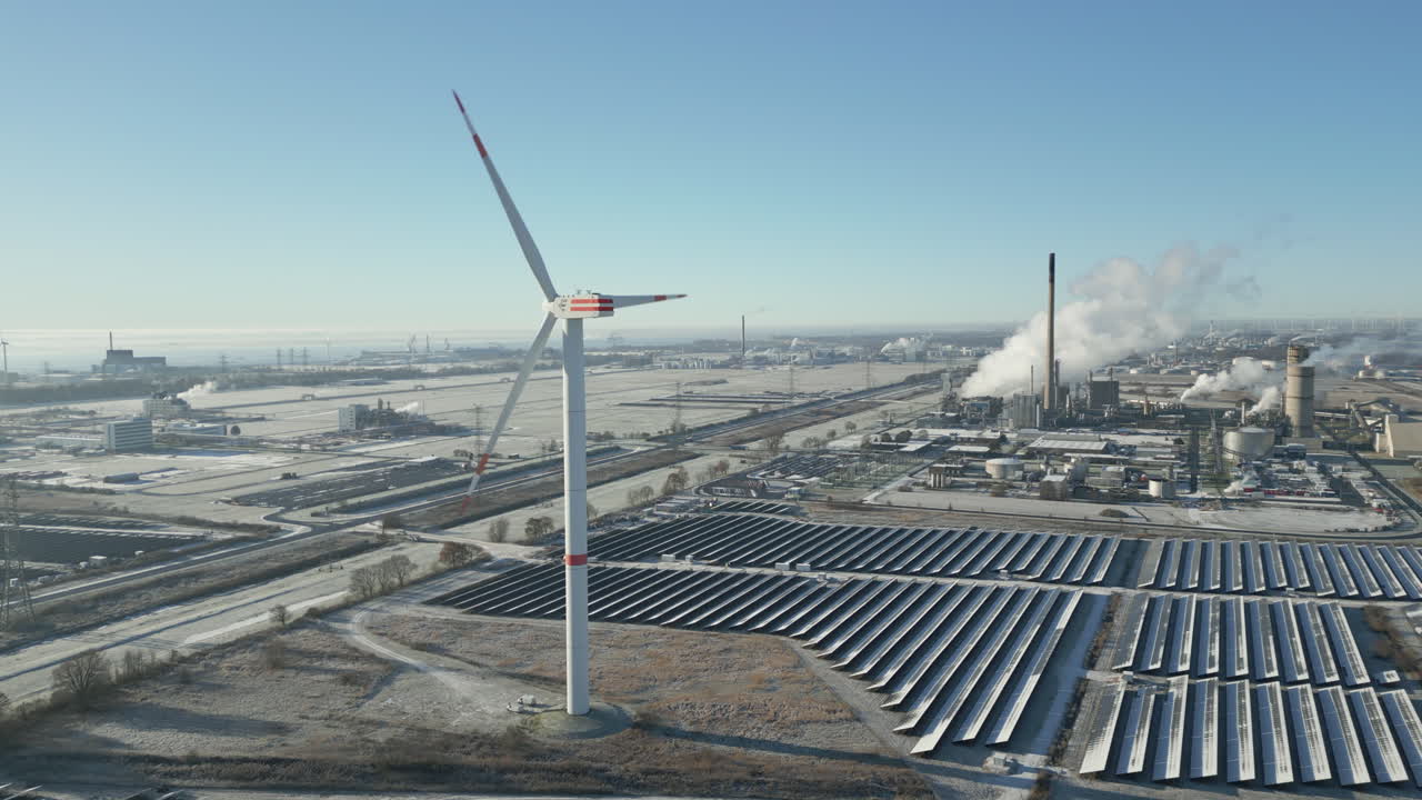Wind turbine spinning in frosty landscape, smoking industrial plant, solar panels, slow moving forward, aerial drone shot