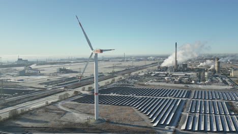 Wind-turbine-spinning-in-frosty-landscape,-smoking-industrial-plant,-solar-panels,-slow-moving-forward,-aerial-drone-shot