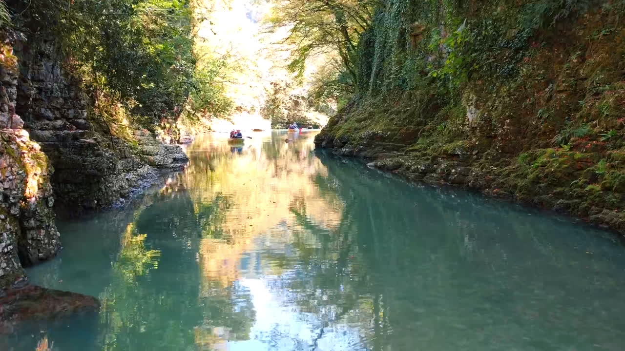 A group of people in a small boat move slowly along a narrow waterway surrounded by high rock walls and lush green trees. The sun shines brightly and reflects on the water