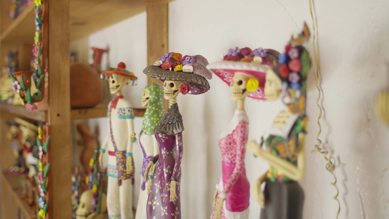 Statuette decorative di Catrina esposte su scaffali all'interno di un laboratorio artigianale, vestiti sui toni del bianco pastello e del viola rosato, profondità di campo ridotta.