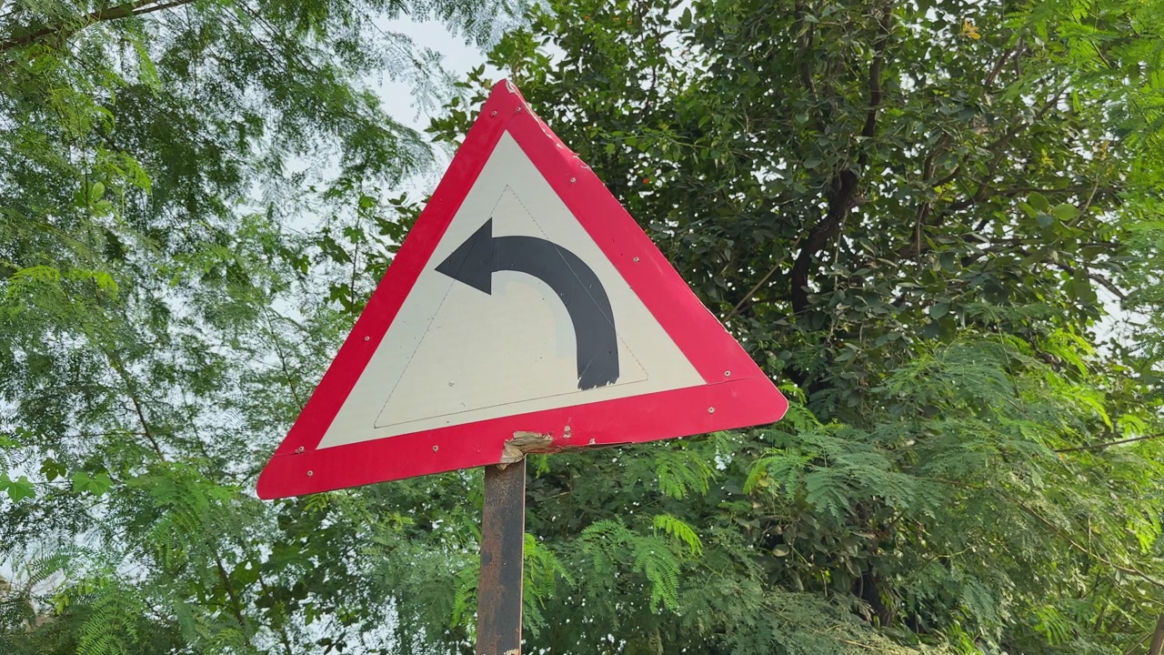 A triangular red-bordered traffic sign indicating a left turn, captured with a circling camera move against a backdrop of green trees.